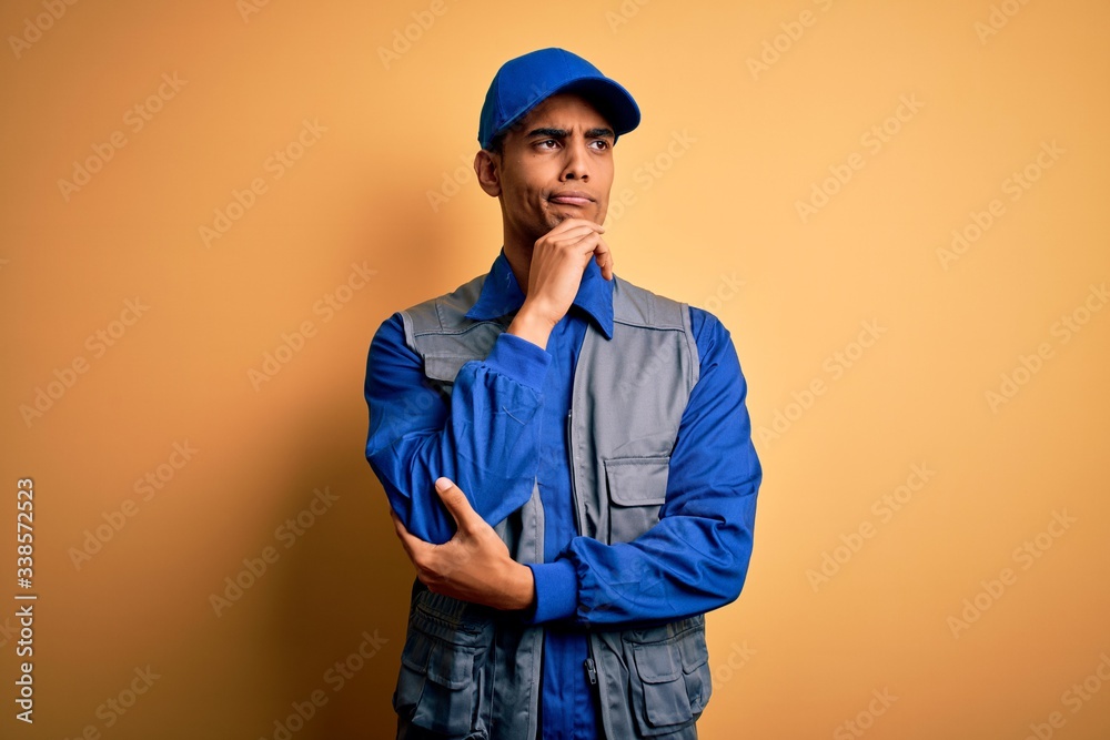 Young handsome african american handyman wearing worker uniform and cap with hand on chin thinking about question, pensive expression. Smiling with thoughtful face. Doubt concept.