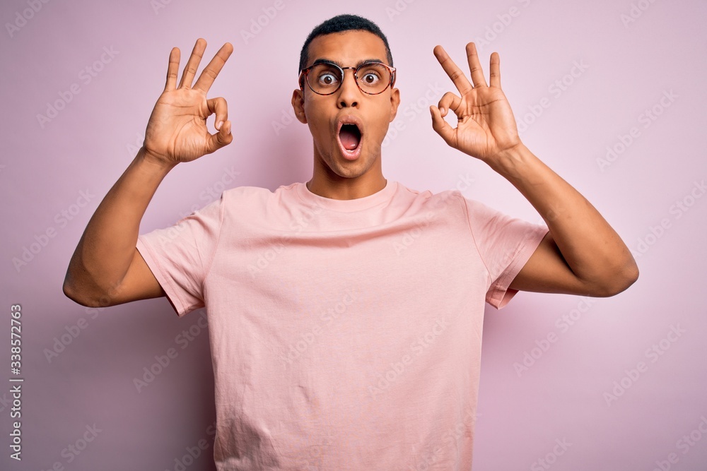 Naklejka premium Handsome african american man wearing casual t-shirt and glasses over pink background looking surprised and shocked doing ok approval symbol with fingers. Crazy expression