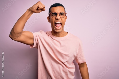 Handsome african american man wearing casual t-shirt and glasses over pink background angry and mad raising fist frustrated and furious while shouting with anger. Rage and aggressive concept.