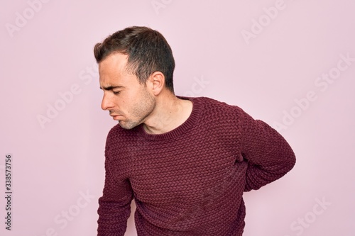 Young handsome man with blue eyes wearing casual sweater standing over pink background Suffering of backache, touching back with hand, muscular pain