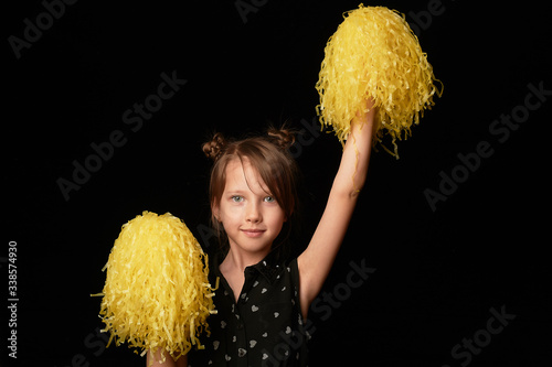 A girl of 6 years, on a black background. A cheerleader with yellow pumpons. Sport and energy.