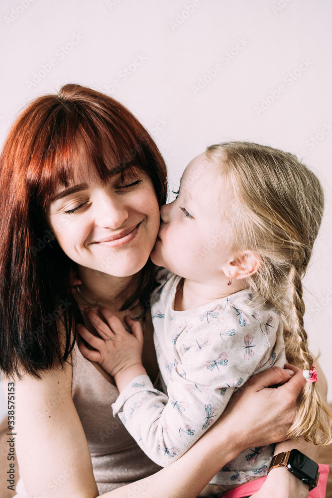 A little blond child gently hugs his brunette mom with handles and kisses her cheek. Joyful mom smiles with closed eyes. Light background. Mother's day concept. Mom and daughter relationship A little blond child gently hugs his brunette mom with handles and kisses her cheek. Joyful mom smiles with closed eyes. Light background. Mother's day concept. Mom and daughter relationship