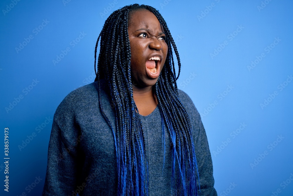 African american plus size woman with braids wearing casual sweater over blue background angry and mad screaming frustrated and furious, shouting with anger. Rage and aggressive concept. African american plus size woman with braids wearing casual sweater over blue background angry and mad screaming frustrated and furious, shouting with anger. Rage and aggressive concept.