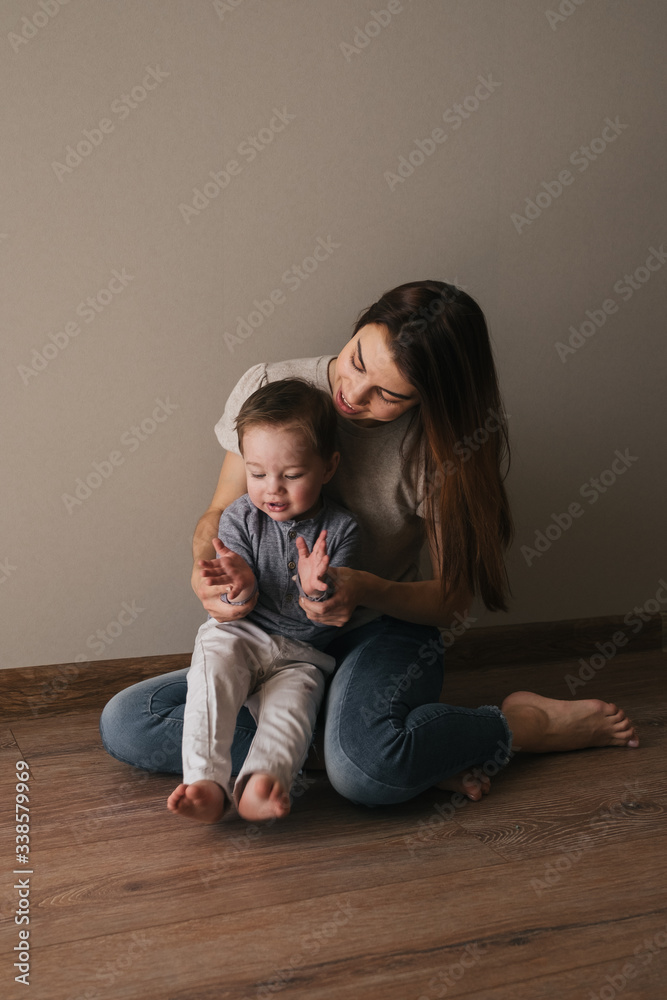 Young beautiful mother plays with her son in a sunny room. Silit with a child on the floor on the carpet, reading a book, playing with toys, tickling, laughing. Quarantine time with your family
