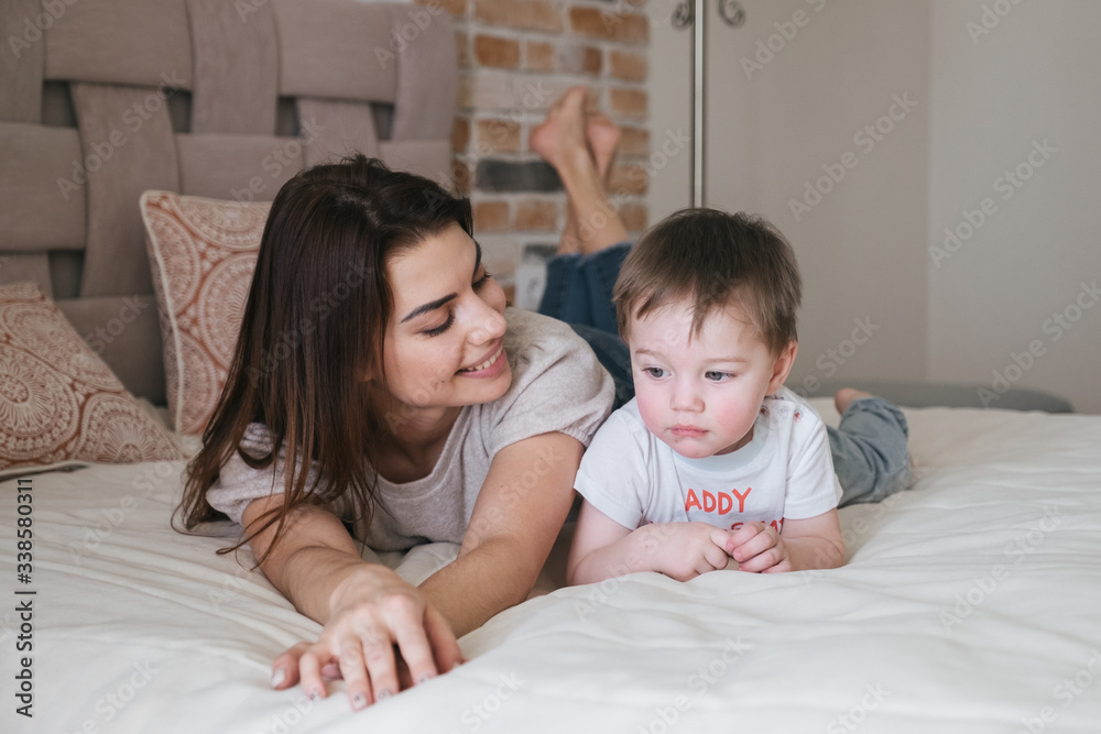 Young beautiful mother plays with her son in a sunny bedroom. Lying on the bed, hugging, tickling, laughing. Quarantine time with your family at home indoors. Love and tender Young beautiful mother plays with her son in a sunny bedroom. Lying on the bed, hugging, tickling, laughing. Quarantine time with your family at home indoors. Love and tender