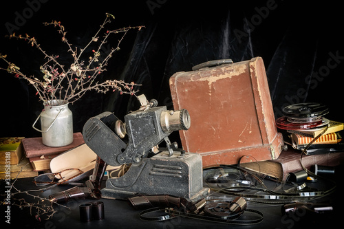 Fototapeta Naklejka Na Ścianę i Meble -  Old vintage projector for viewing filmstrips on background of wooden packaging box, reels with film strip, piles of old books and dried flowers. Nostalgia concept