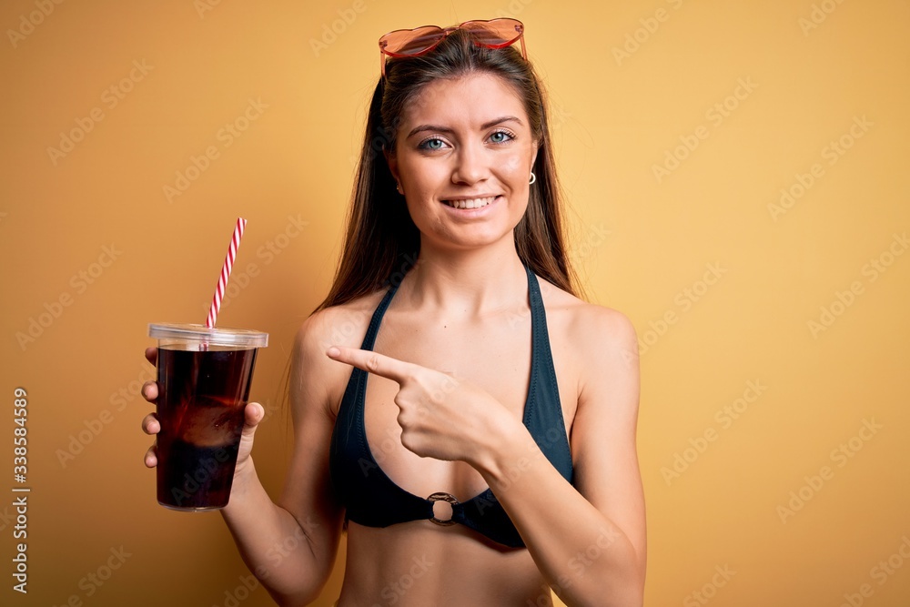 Young beautiful woman with blue eyes on vacation wearing bikini drinking cola beverage very happy pointing with hand and finger Young beautiful woman with blue eyes on vacation wearing bikini drinking cola beverage very happy pointing with hand and finger