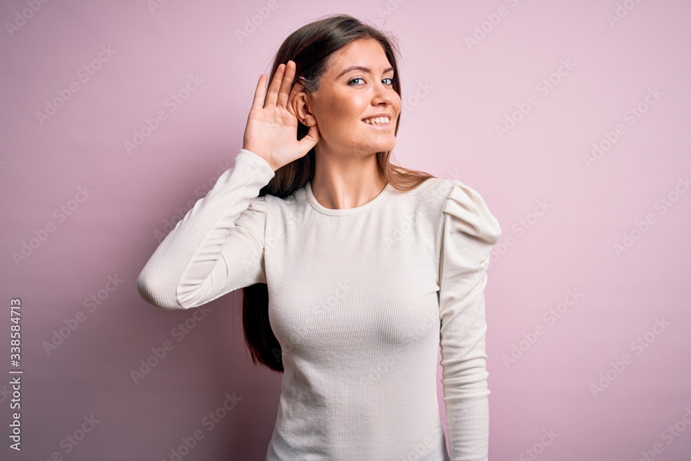 Young beautiful woman with blue eyes wearing casual white t-shirt over pink background smiling with hand over ear listening an hearing to rumor or gossip. Deafness concept. Young beautiful woman with blue eyes wearing casual white t-shirt over pink background smiling with hand over ear listening an hearing to rumor or gossip. Deafness concept.