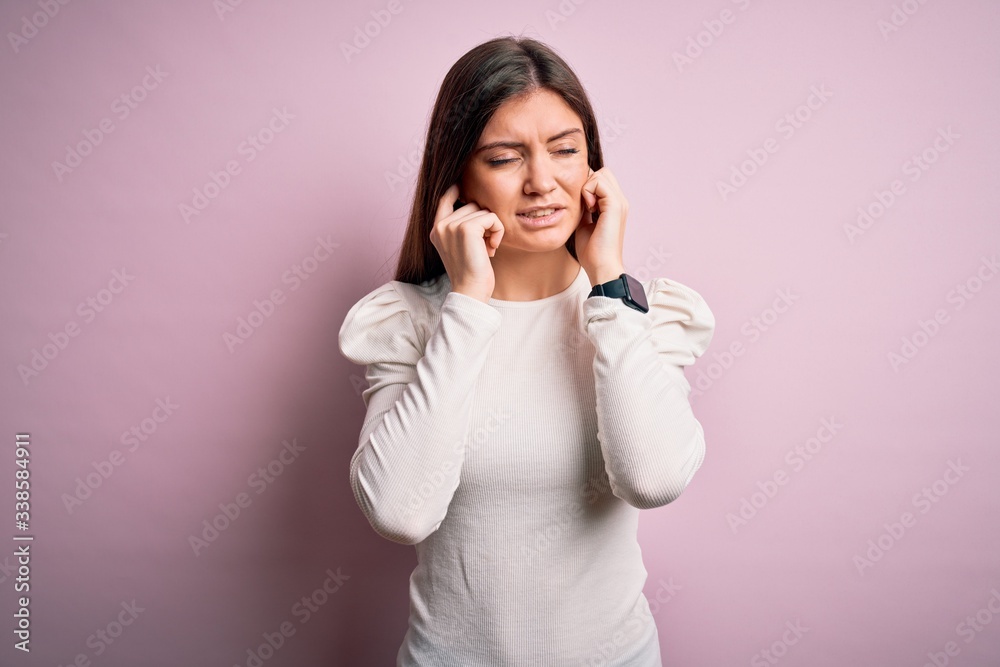 Young beautiful woman with blue eyes wearing casual white t-shirt over pink background covering ears with fingers with annoyed expression for the noise of loud music. Deaf concept. Young beautiful woman with blue eyes wearing casual white t-shirt over pink background covering ears with fingers with annoyed expression for the noise of loud music. Deaf concept.