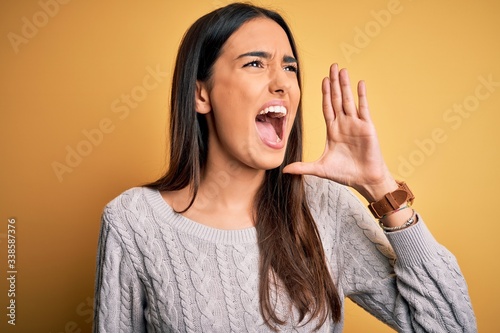 Young beautiful brunette woman wearing white casual sweater over yellow background shouting and screaming loud to side with hand on mouth. Communication concept.