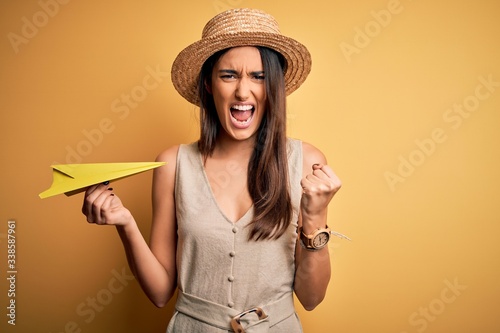 Young beautiful brunette woman wearing hat holding paper airplane over yellow background annoyed and frustrated shouting with anger  crazy and yelling with raised hand  anger concept