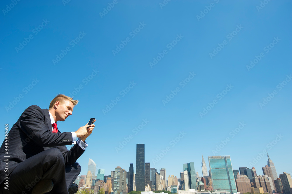 Cheerful businessman sitting outdoors using his mobile phone in front of the city skyline under clear blue sky copy space Cheerful businessman sitting outdoors using his mobile phone in front of the city skyline under clear blue sky copy space