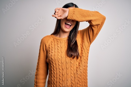 Young beautiful brunette woman wearing casual sweater over isolated white background covering eyes with arm smiling cheerful and funny. Blind concept.