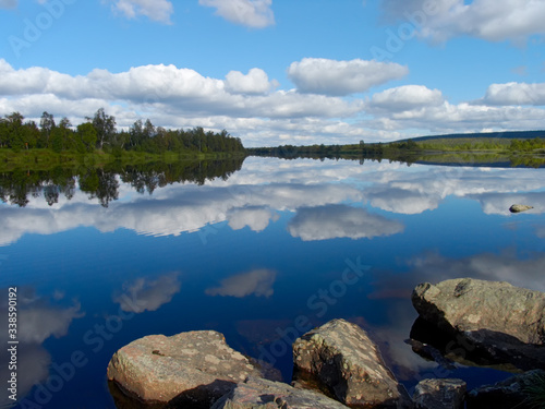 North. A river going into the distance. A blue sky with clouds is reflected in the water. In the foreground there are three stones. In the distance you can see the shore, covered with trees.