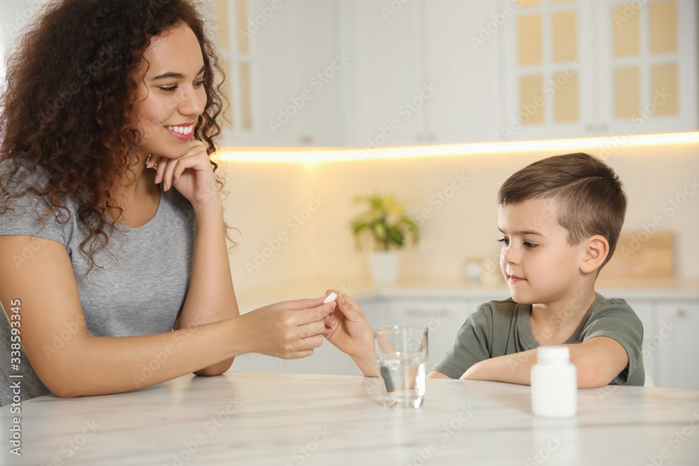 African-American woman giving vitamin pill to little boy in kitchen African-American woman giving vitamin pill to little boy in kitchen