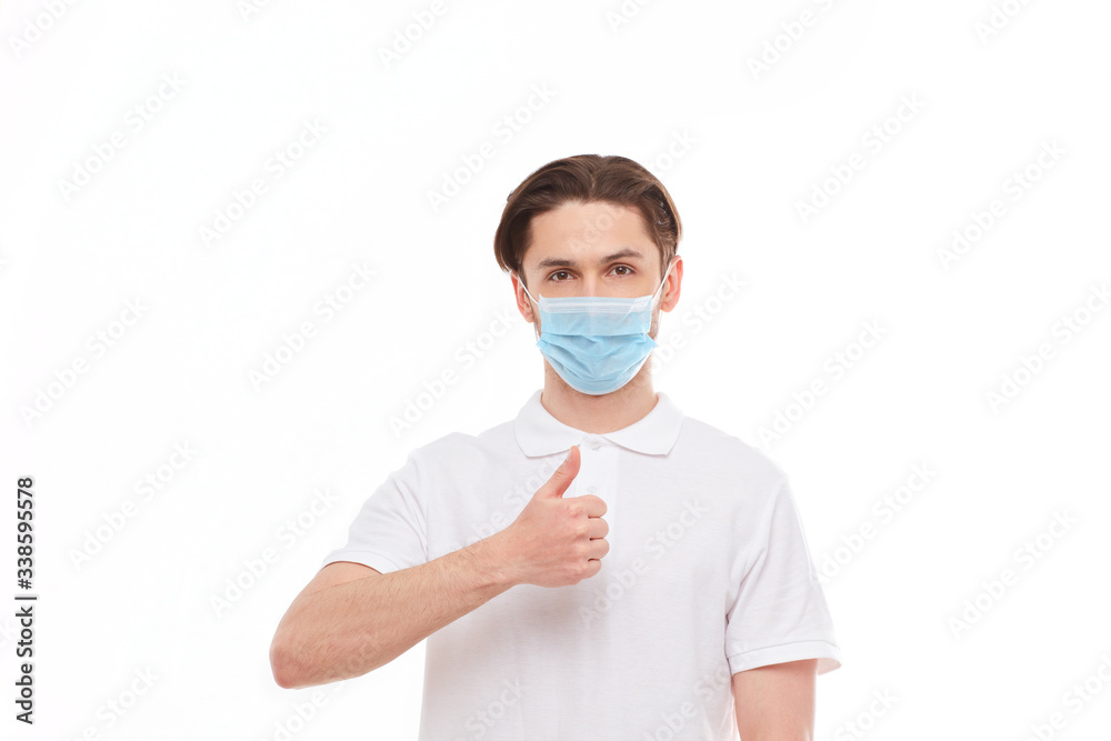 A man in a white T-shirt of European appearance in a medical mask, shows a thumb up. In the mask you are protected from covid 19 viruses.