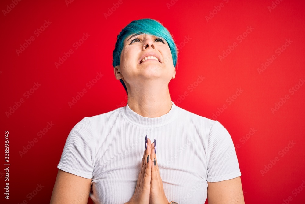 Young beautiful woman with blue fashion hair wearing casual t-shirt over red background begging and praying with hands together with hope expression on face very emotional and worried. Begging. Young beautiful woman with blue fashion hair wearing casual t-shirt over red background begging and praying with hands together with hope expression on face very emotional and worried. Begging.