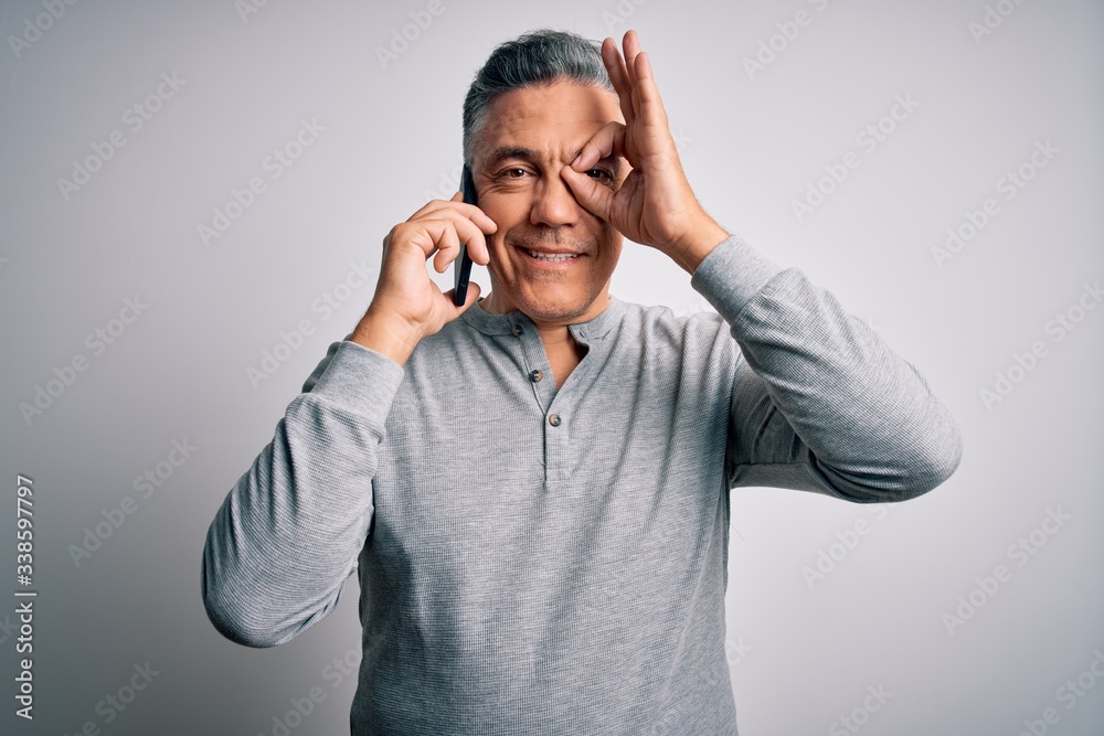 Middle age handsome grey-haired man having conversation talking on the smartphone with happy face smiling doing ok sign with hand on eye looking through fingers Middle age handsome grey-haired man having conversation talking on the smartphone with happy face smiling doing ok sign with hand on eye looking through fingers