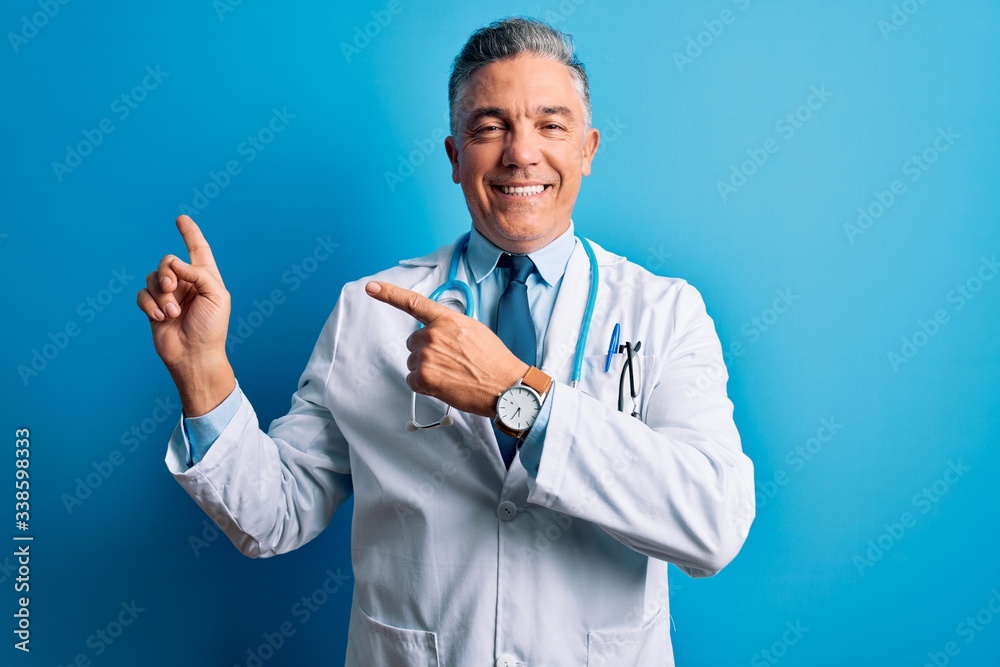 Middle age handsome grey-haired doctor man wearing coat and blue stethoscope smiling and looking at the camera pointing with two hands and fingers to the side. Middle age handsome grey-haired doctor man wearing coat and blue stethoscope smiling and looking at the camera pointing with two hands and fingers to the side.
