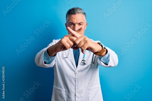 Middle age handsome grey-haired doctor man wearing coat and blue stethoscope Rejection expression crossing fingers doing negative sign