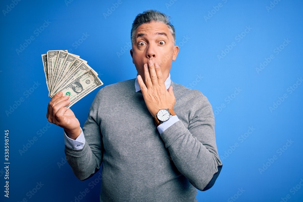 Middle age handsome grey-haired man holding bunch of dollars over blue background cover mouth with hand shocked with shame for mistake, expression of fear, scared in silence, secret concept Middle age handsome grey-haired man holding bunch of dollars over blue background cover mouth with hand shocked with shame for mistake, expression of fear, scared in silence, secret concept
