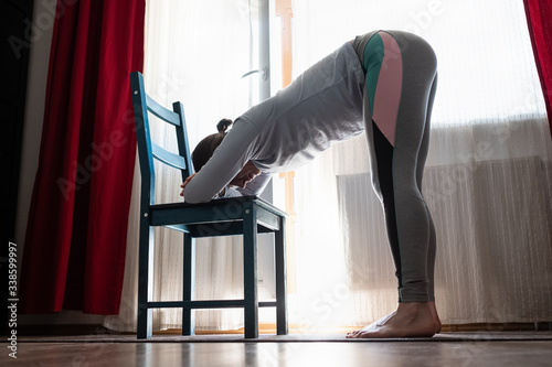 Young woman practicing yoga, doing head to knees, uttanasana exercise, Standing forward bend pose, using chair at the living room photo