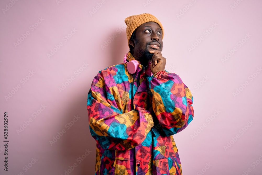 Young handsome african american man wearing colorful coat and cap over pink background with hand on chin thinking about question, pensive expression. Smiling with thoughtful face. Doubt concept.