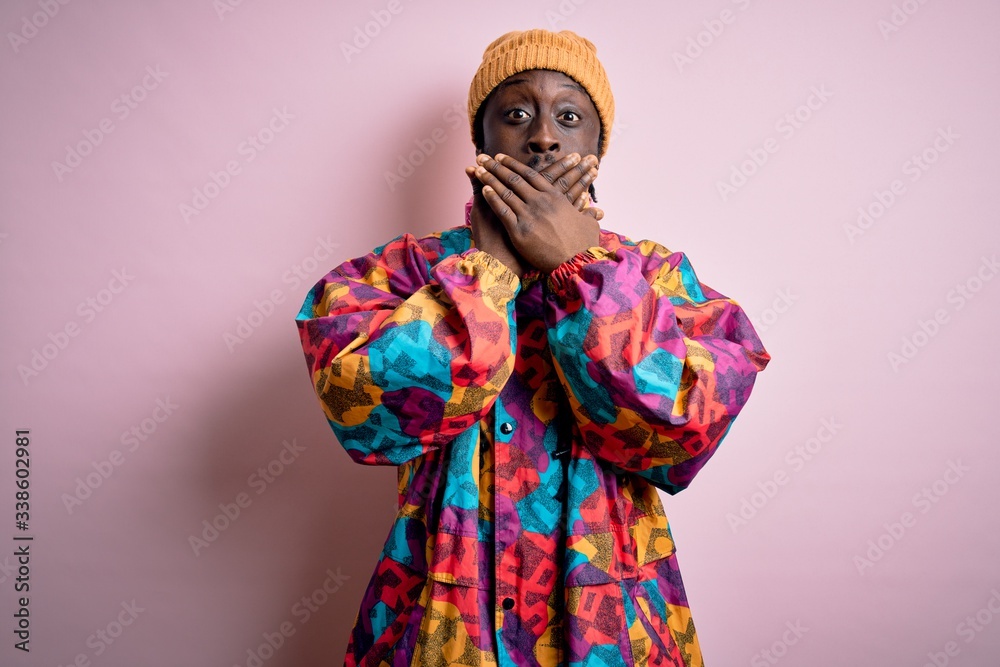 Young handsome african american man wearing colorful coat and cap over pink background shocked covering mouth with hands for mistake. Secret concept.