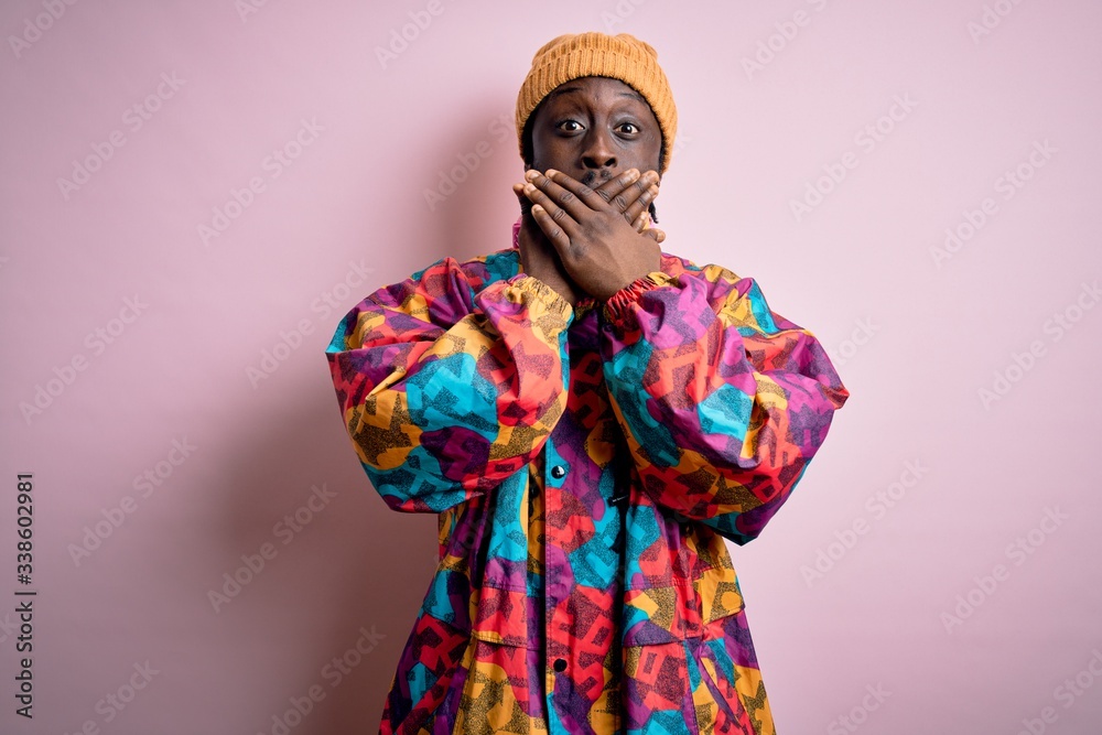 Young handsome african american man wearing colorful coat and cap over pink background shocked covering mouth with hands for mistake. Secret concept. Young handsome african american man wearing colorful coat and cap over pink background shocked covering mouth with hands for mistake. Secret concept.