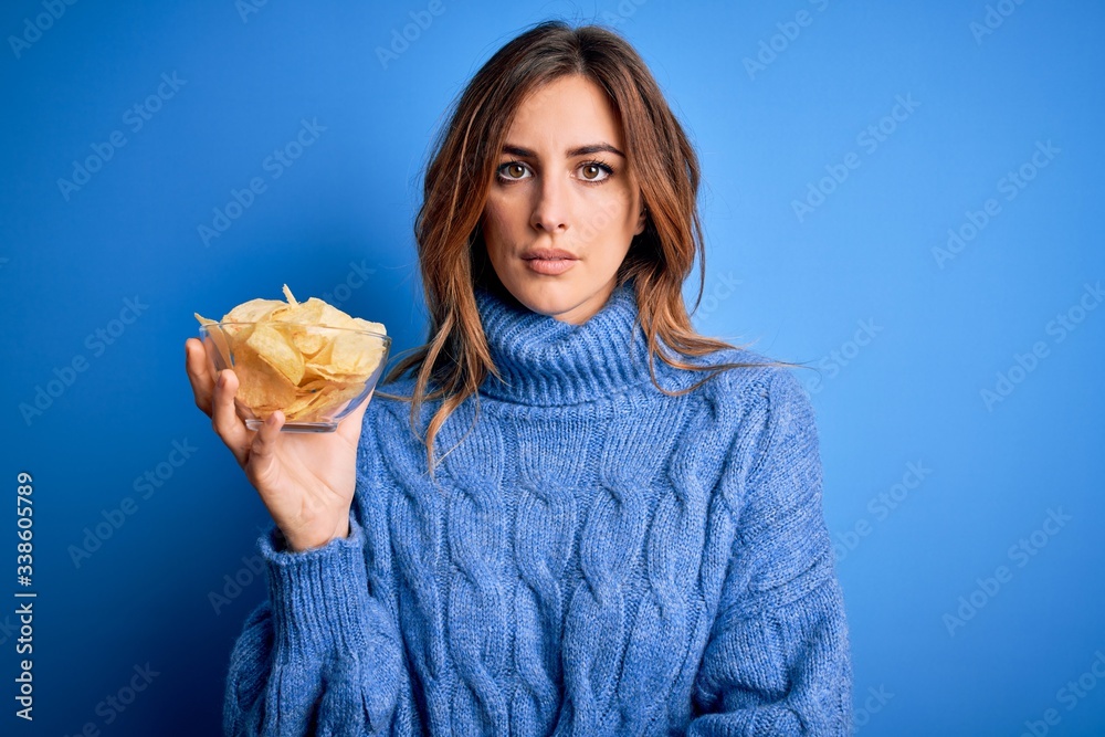 Young beautiful brunette woman holding bowl with potato chips over blue background with a confident expression on smart face thinking serious Young beautiful brunette woman holding bowl with potato chips over blue background with a confident expression on smart face thinking serious