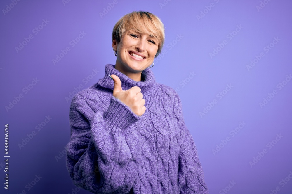 Young blonde woman with short hair wearing winter turtleneck sweater over purple background doing happy thumbs up gesture with hand. Approving expression looking at the camera showing success. Young blonde woman with short hair wearing winter turtleneck sweater over purple background doing happy thumbs up gesture with hand. Approving expression looking at the camera showing success.