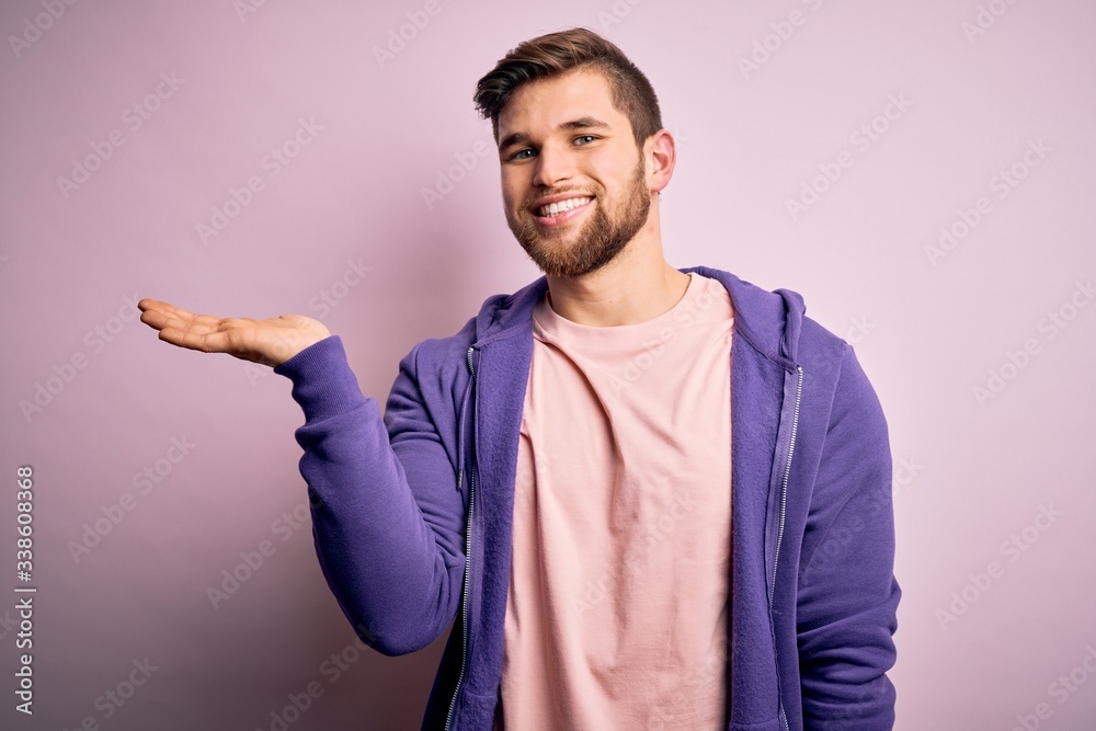 Young blond man with beard and blue eyes wearing purple sweatshirt over pink background smiling cheerful presenting and pointing with palm of hand looking at the camera. Young blond man with beard and blue eyes wearing purple sweatshirt over pink background smiling cheerful presenting and pointing with palm of hand looking at the camera.