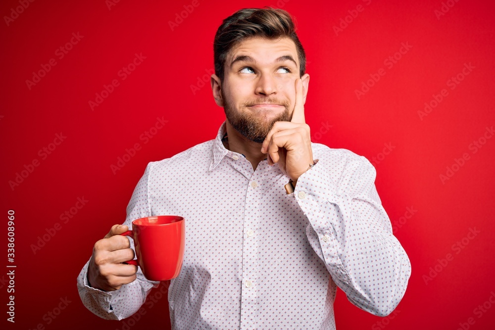 Young blond businessman with beard and blue eyes drinking red cup of coffee serious face thinking about question, very confused idea Young blond businessman with beard and blue eyes drinking red cup of coffee serious face thinking about question, very confused idea
