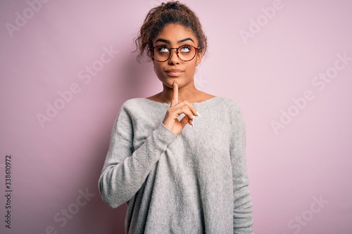 Young beautiful african american girl wearing sweater and glasses over pink background Thinking concentrated about doubt with finger on chin and looking up wondering