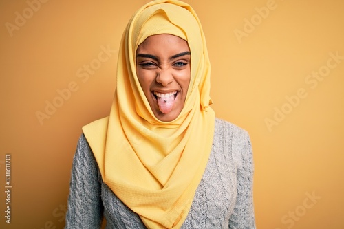 Young beautiful african american girl wearing muslim hijab over isolated yellow background sticking tongue out happy with funny expression. Emotion concept. © Krakenimages.com