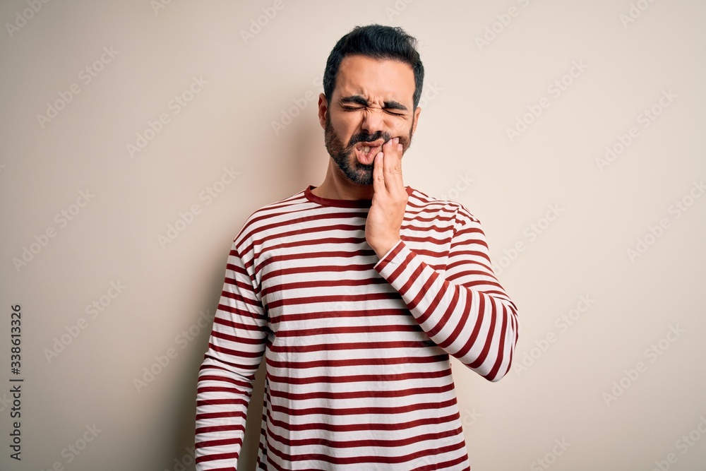 Young handsome man with beard wearing casual striped t-shirt standing over white background touching mouth with hand with painful expression because of toothache or dental illness on teeth. Dentist
