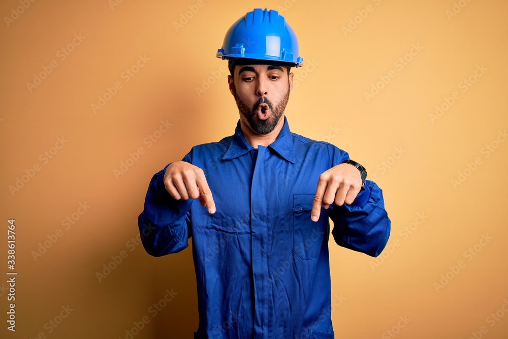 Mechanic man with beard wearing blue uniform and safety helmet over yellow background Pointing down with fingers showing advertisement, surprised face and open mouth