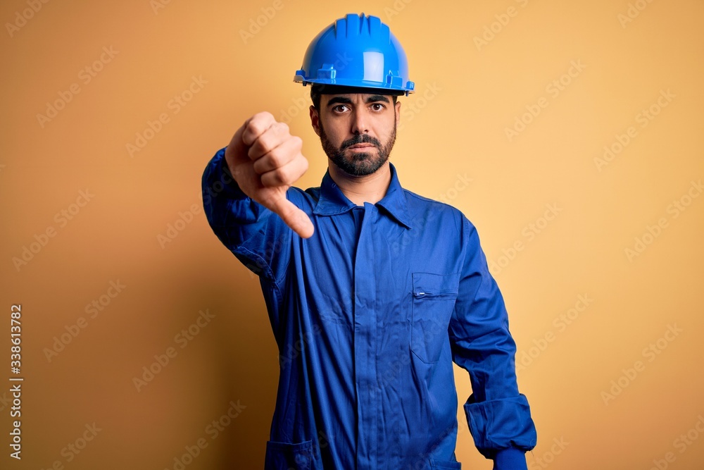Mechanic man with beard wearing blue uniform and safety helmet over yellow background looking unhappy and angry showing rejection and negative with thumbs down gesture. Bad expression.