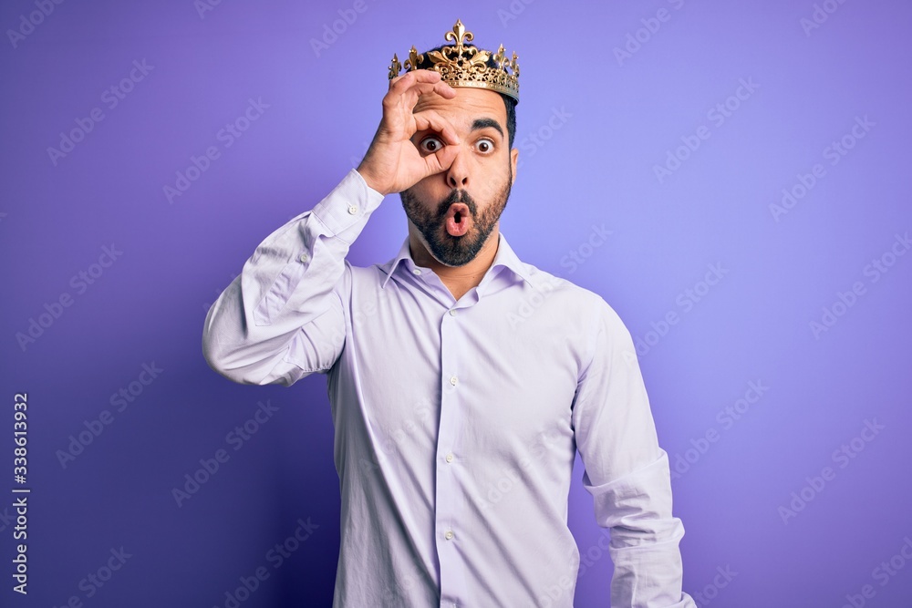 Young handsome man with beard wearing golden crown of king over purple background doing ok gesture shocked with surprised face, eye looking through fingers. Unbelieving expression.