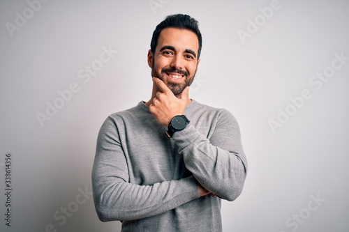Young handsome man with beard wearing casual sweater standing over white background looking confident at the camera smiling with crossed arms and hand raised on chin. Thinking positive.