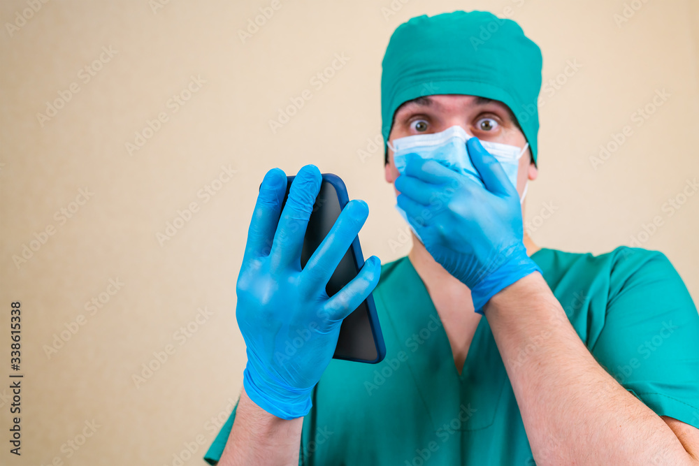 Doctor in protective medical mask shocked looking news in his mobile phone in hand isolated on smooth background with copy space for text. Selective focus. Doctor in protective medical mask shocked looking news in his mobile phone in hand isolated on smooth background with copy space for text. Selective focus.