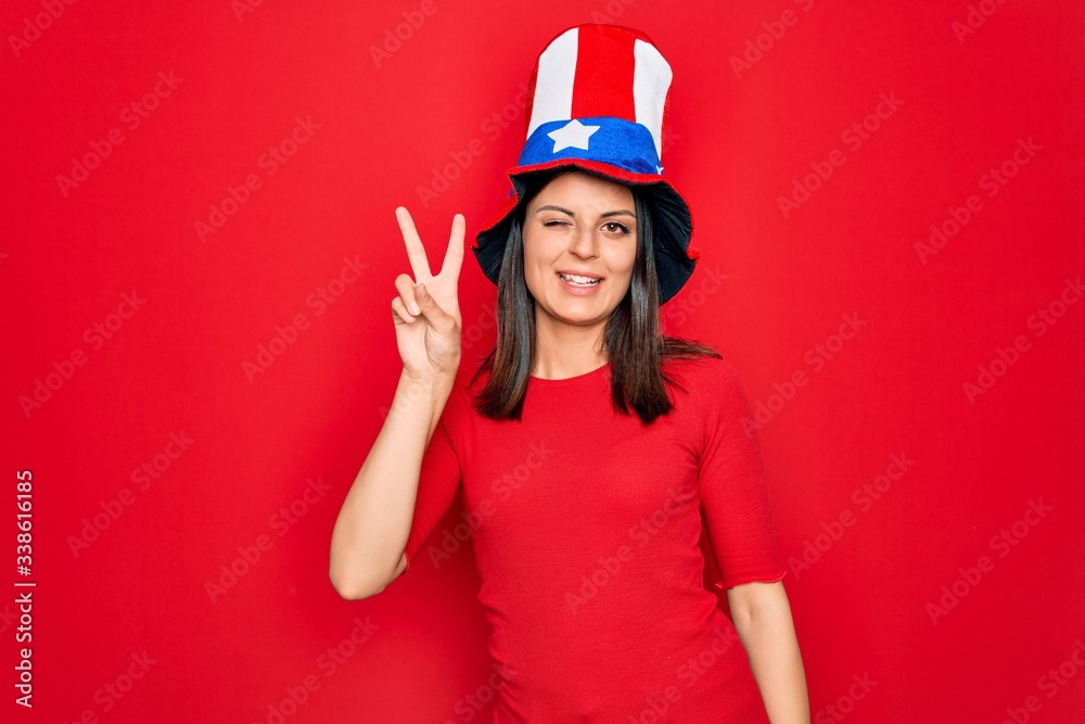 Young beautiful brunette woman wearing united states hat celebrating independence day smiling with happy face winking at the camera doing victory sign with fingers. Number two.