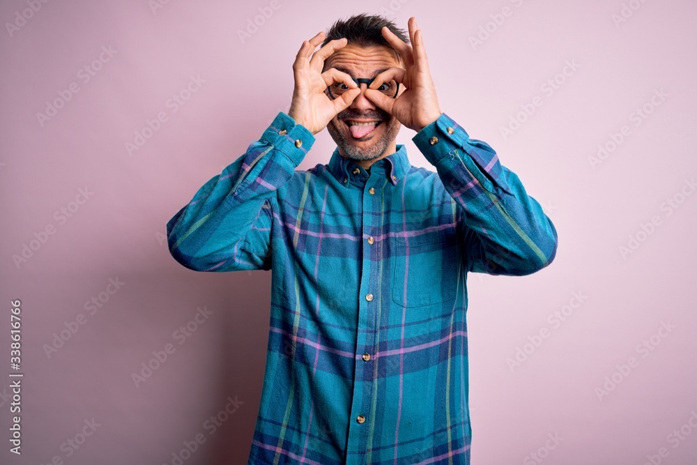 Young handsome man wearing casual shirt and glasses standing over isolated pink background doing ok gesture like binoculars sticking tongue out, eyes looking through fingers. Crazy expression. Young handsome man wearing casual shirt and glasses standing over isolated pink background doing ok gesture like binoculars sticking tongue out, eyes looking through fingers. Crazy expression.