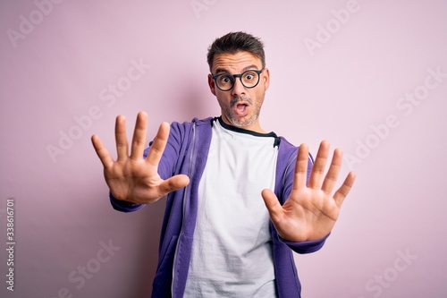 Young handsome man wearing purple sweatshirt and glasses standing over pink background doing stop gesture with hands palms, angry and frustration expression