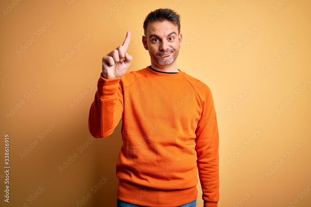 Young handsome man wearing orange casual sweater standing over isolated yellow background showing and pointing up with finger number one while smiling confident and happy.
