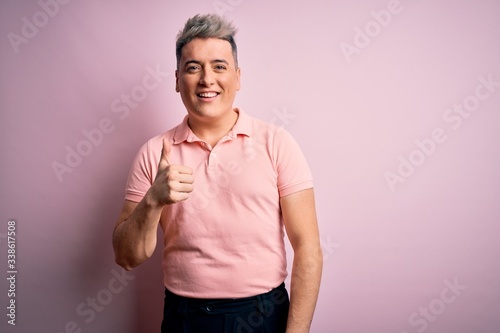 Young handsome modern man wearing casual pink t-shirt over isolated background doing happy thumbs up gesture with hand. Approving expression looking at the camera showing success.