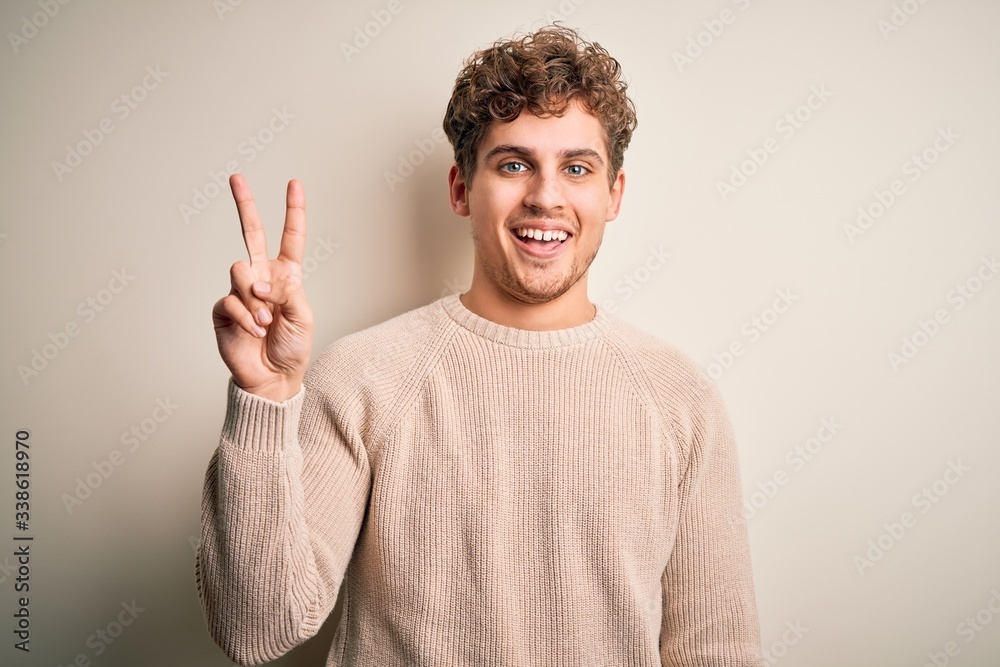 Young blond handsome man with curly hair wearing casual sweater over white background showing and pointing up with fingers number two while smiling confident and happy.