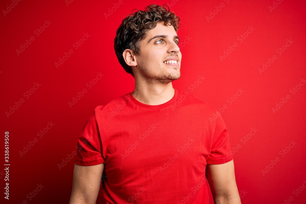 Young blond handsome man with curly hair wearing casual t-shirt over red background looking away to side with smile on face, natural expression. Laughing confident. Young blond handsome man with curly hair wearing casual t-shirt over red background looking away to side with smile on face, natural expression. Laughing confident.