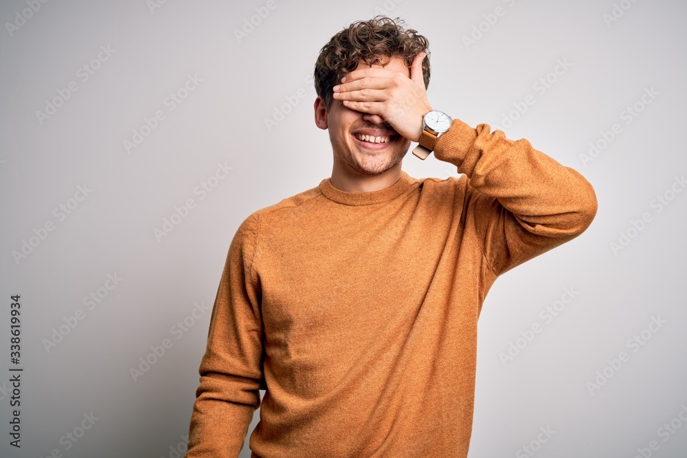 Young blond handsome man with curly hair wearing casual sweater over white background smiling and laughing with hand on face covering eyes for surprise. Blind concept. Young blond handsome man with curly hair wearing casual sweater over white background smiling and laughing with hand on face covering eyes for surprise. Blind concept.
