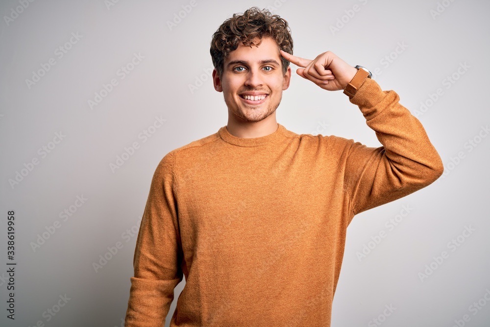 Young blond handsome man with curly hair wearing casual sweater over white background Smiling pointing to head with one finger, great idea or thought, good memory Young blond handsome man with curly hair wearing casual sweater over white background Smiling pointing to head with one finger, great idea or thought, good memory
